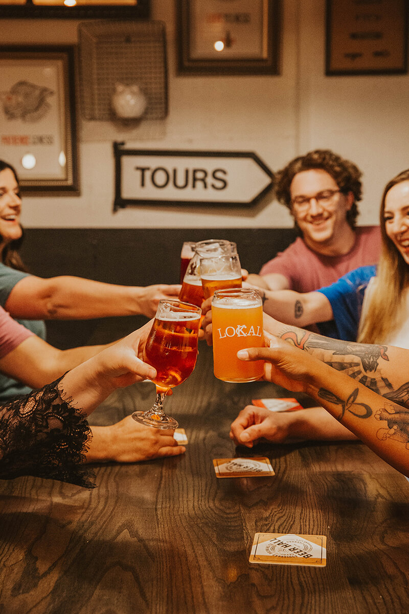 A group of people around a wooden table clinking glasses filled with various beverages. The setting is casual with framed pictures and a "TOURS" sign on the wall in the background. The table has playing cards scattered on it, and everyone is smiling and convivial.
